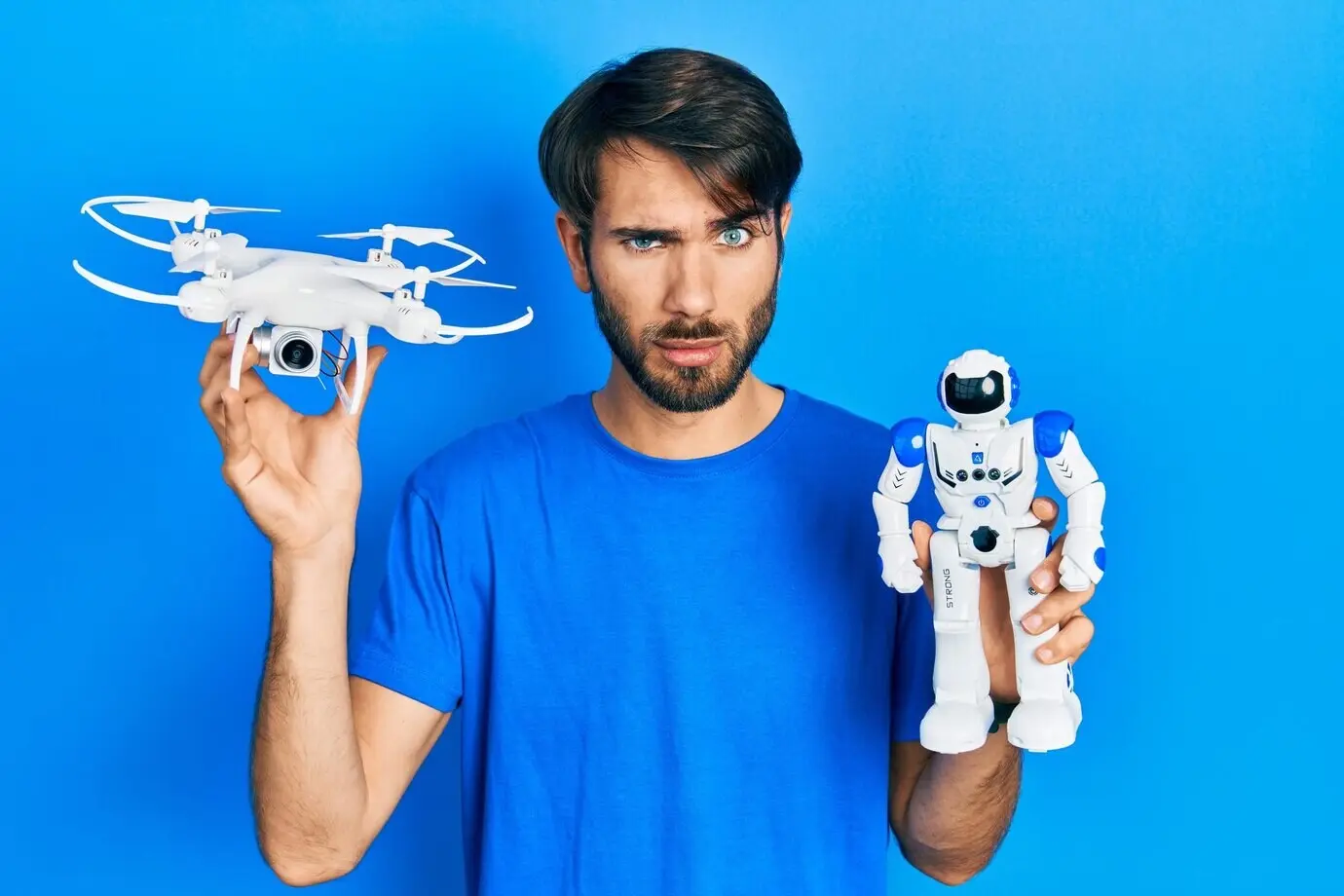 Junger hispanischer Mann hält eine Drohne und ein Roboterspielzeug, wirkt skeptisch und nervös, runzelt die Stirn und ist wegen eines Problems verärgert. Negative Person.
